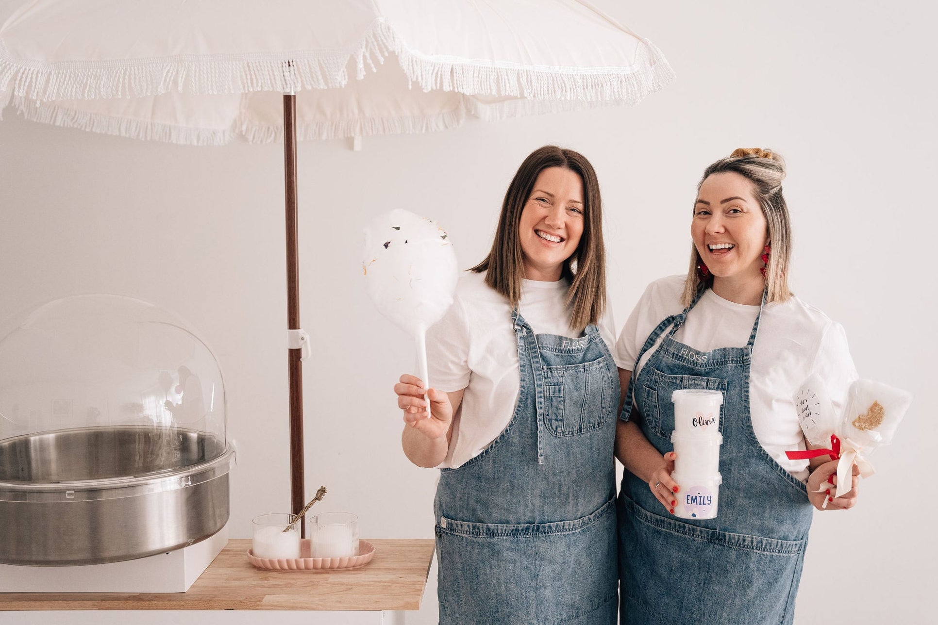 Twins Emma and Claire in denim aprons holding a white fairy floss stick and a tubs of personalised fairy floss