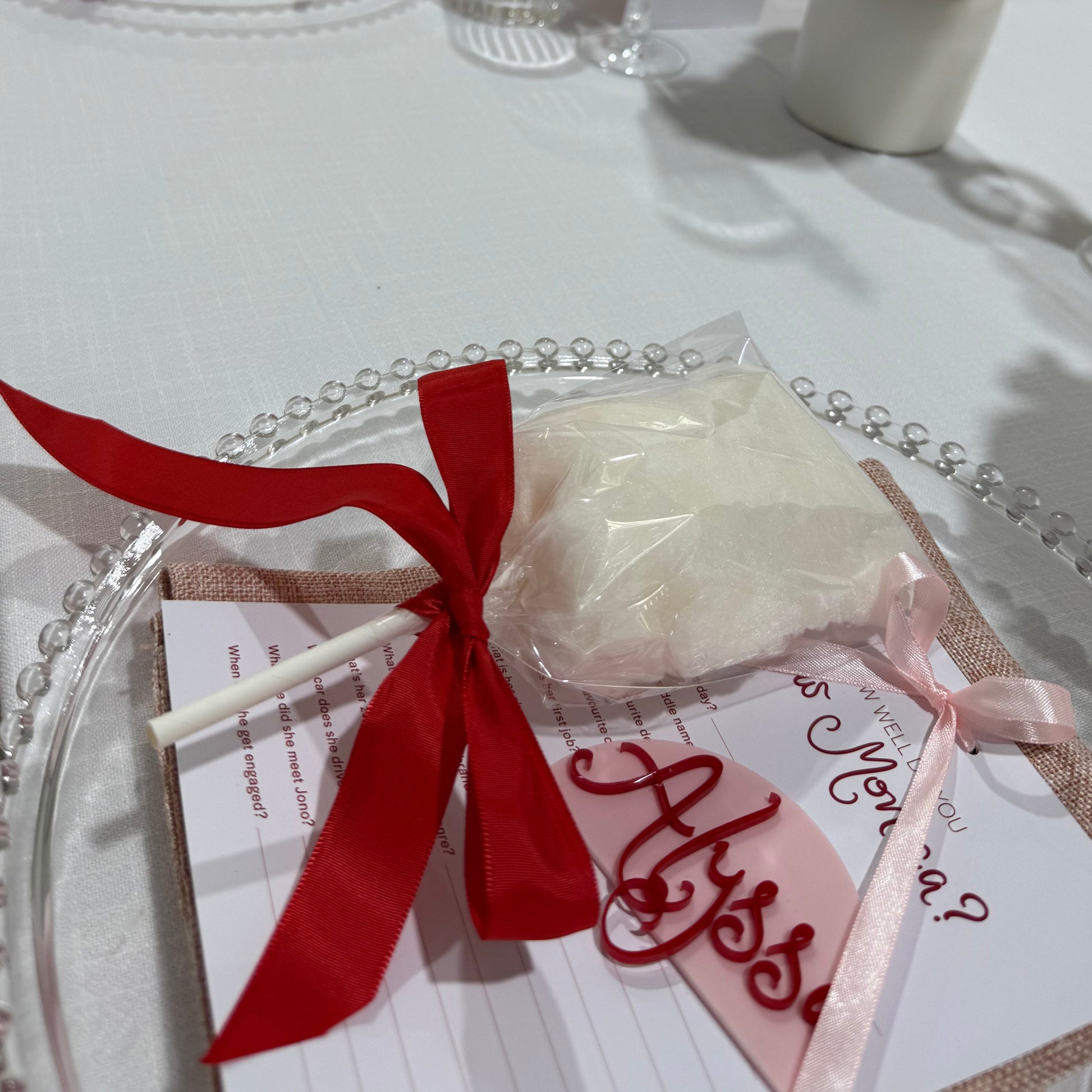A close-up of a fairy floss stick with a red ribbon on a plate, part of a bundle, with blurred background suggesting a festive or event setting.