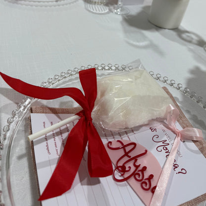 A close-up of a fairy floss stick with a red ribbon on a plate, part of a bundle, with blurred background suggesting a festive or event setting.